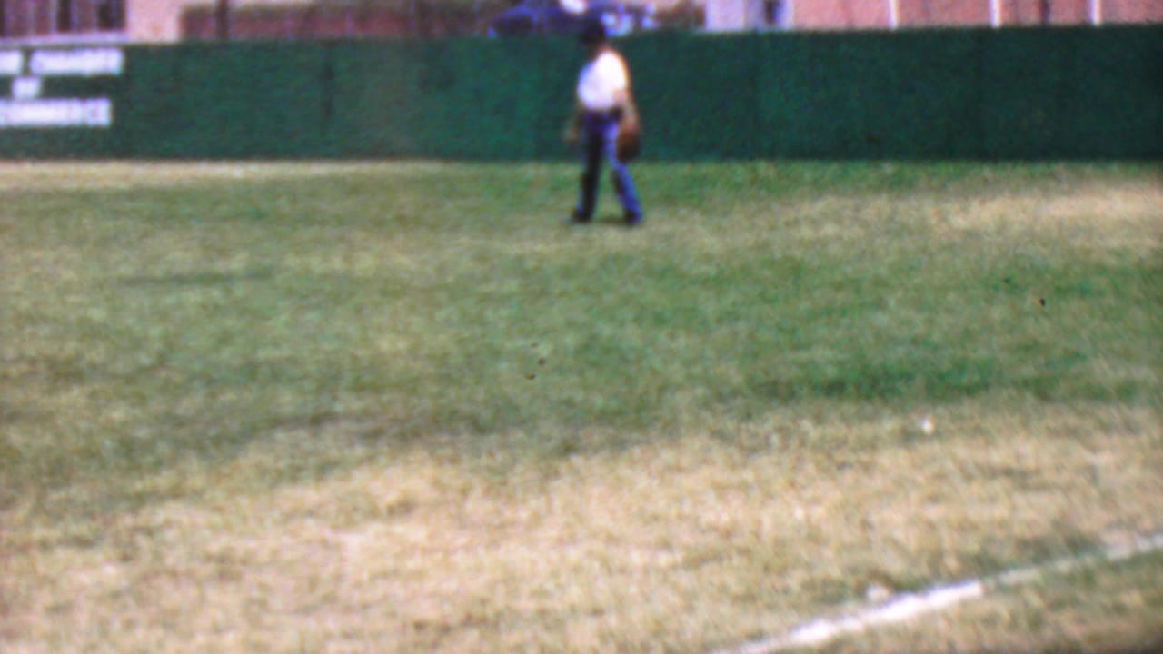 Distant person in a white shirt walking across a grassy outfield near a green wall.