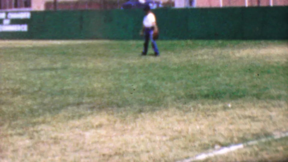 1964 Anaheim Disneyland home-movie frame showing a distant figure in a white shirt and dark pants carrying a glove across a grassy baseball outfield by a green fence.