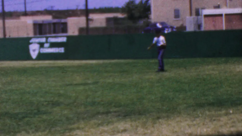 1964 metadata-supported Anaheim Disneyland frame with a distant person holding a baseball glove on the outfield grass, backed by a green wall and Junior Chamber of Commerce signage.