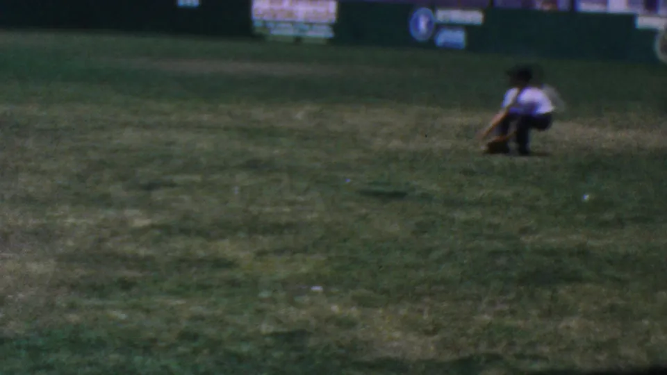 1964 Anaheim home-movie frame showing a baseball player crouched on a grass field, with another distant figure and outfield advertising boards behind him.