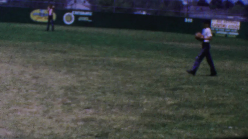 1964 Anaheim 8mm frame showing a baseball player in a light shirt and dark trousers moving across the grass, with outfield signage and another distant figure in the background.