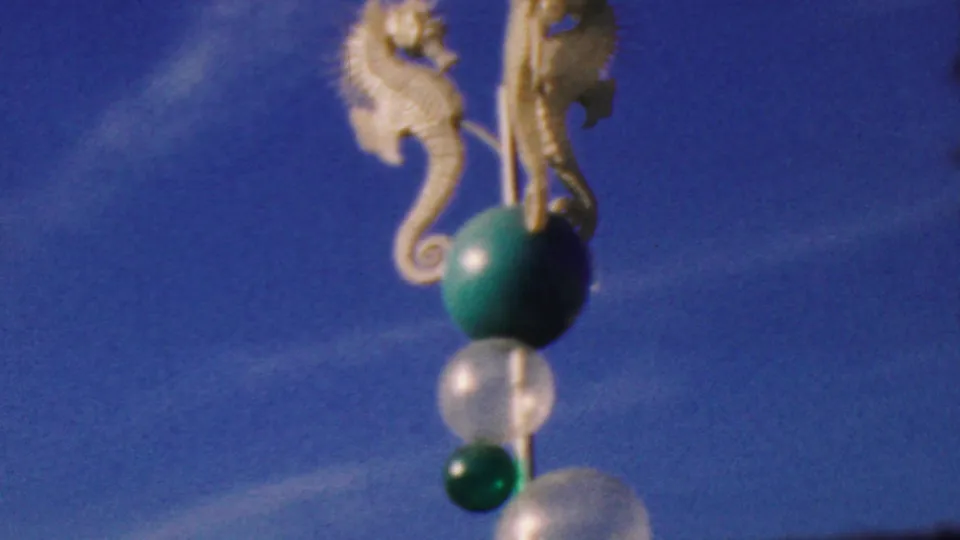 1964 Disneyland home-movie frame showing white seahorse figures beside a decorative pole with green and translucent spheres, seen from below against blue sky.