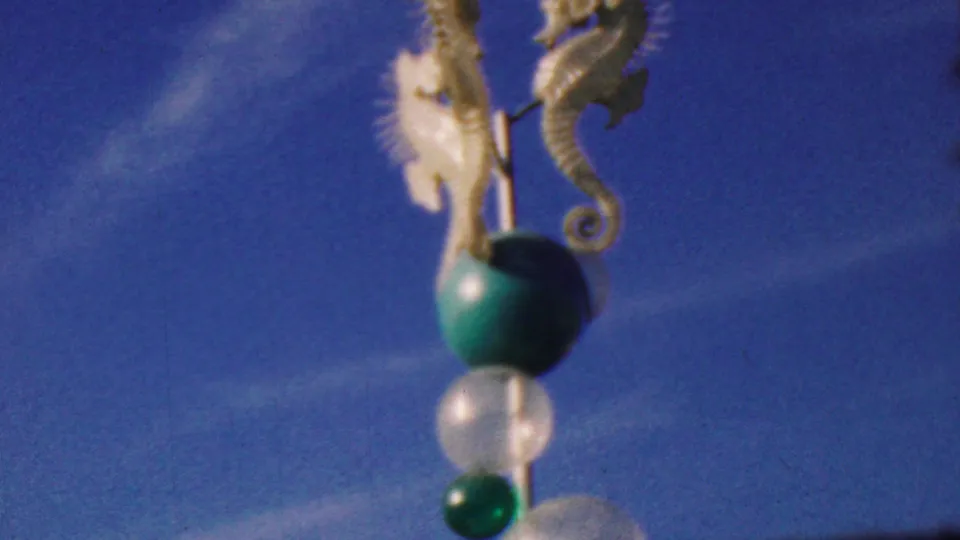 1964 Disneyland home-movie frame showing paired white seahorse ornaments mounted near green and clear spheres on a vertical decorative pole against open sky.