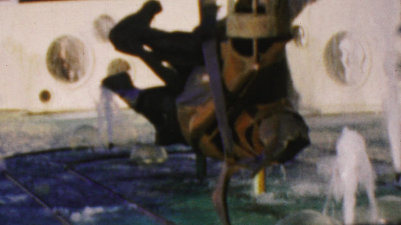 1964 Anaheim 8mm frame showing a dark sheet-metal human-like fountain sculpture with water jets, round wall ornaments, and blue-green water.