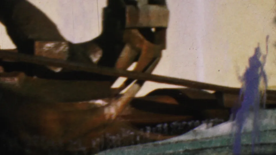 1964 Anaheim home-movie frame with a close angle on dark sheet-metal sculpture parts, a raised water jet, and the curved edge of the fountain basin.