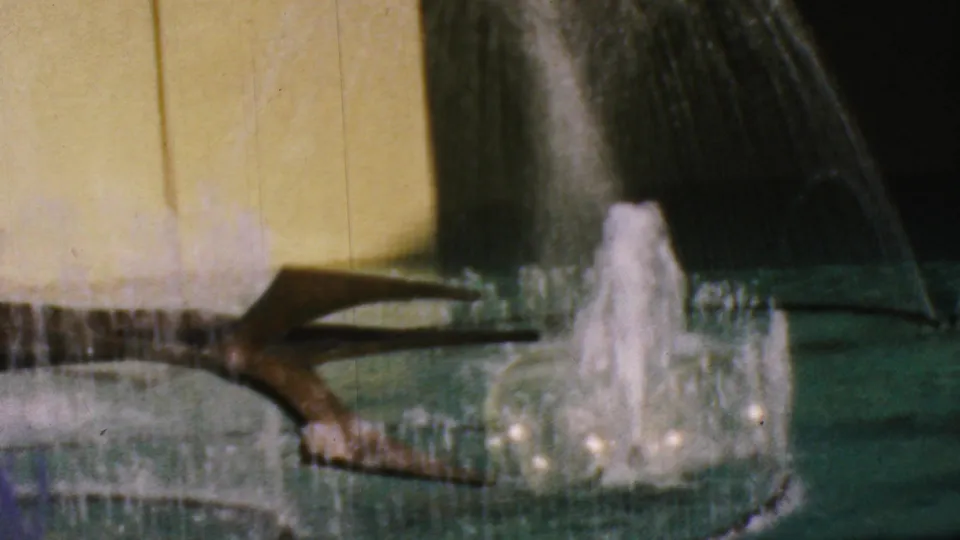 1964 Anaheim Disneyland frame with a white fountain plume, dark arcing fountain element, green basin, and yellow wall backdrop.