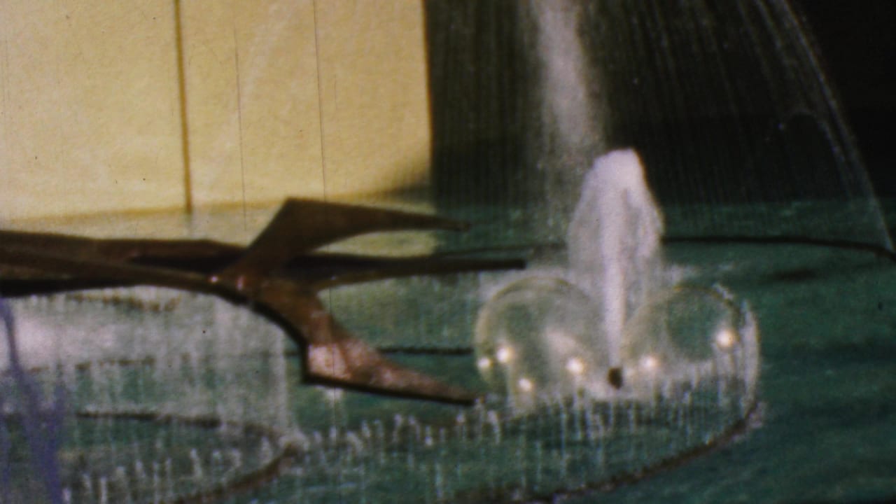 1964 Anaheim Disneyland home-movie frame showing arcing fountain water, a central splash, green pool water, a yellow wall, and a dark sculptural element.