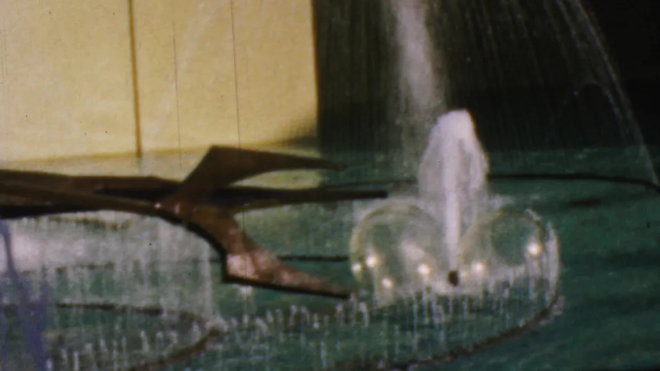 1964 Anaheim Disneyland home-movie frame showing arcing fountain water, a central splash, green pool water, a yellow wall, and a dark sculptural element.
