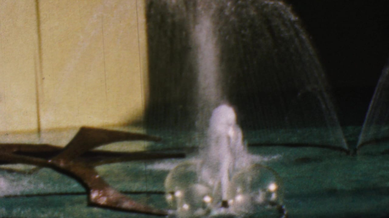 1964 Anaheim Disneyland frame with a close fountain splash, rounded water forms, green pool surface, yellow wall, and dark sculptural feature partly crossing the scene.