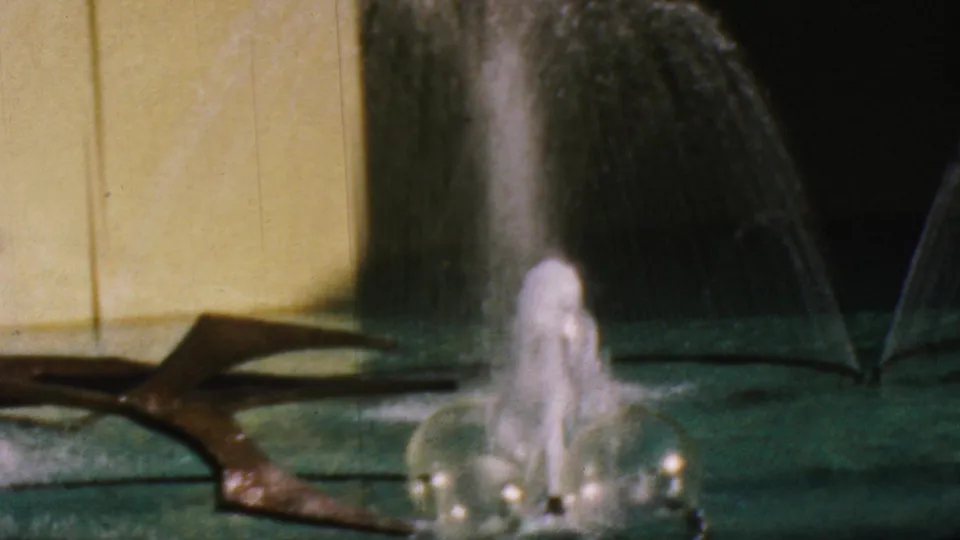 1964 Anaheim Disneyland frame with a close fountain splash, rounded water forms, green pool surface, yellow wall, and dark sculptural feature partly crossing the scene.