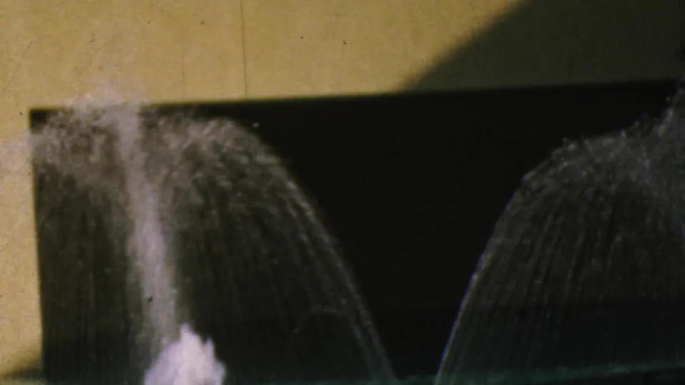 1964 Anaheim Disneyland home-movie frame focused on arcing fountain spray and a white plume against a yellow wall and darkened pool background.
