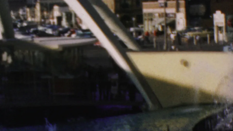 1964 Disneyland visitor footage looking through white diagonal supports toward fountain spray, parked cars, buildings, and small background figures.