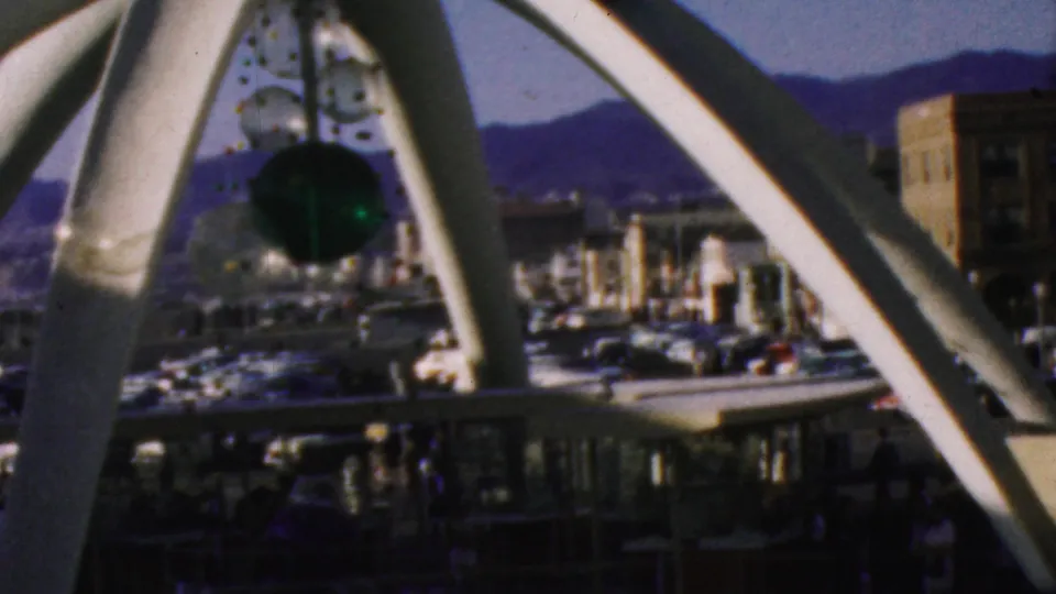Wider 1964 Anaheim Disneyland frame showing a white structural frame, a dark hanging element, fountain area, parked cars, buildings, small visitors, and distant hills.