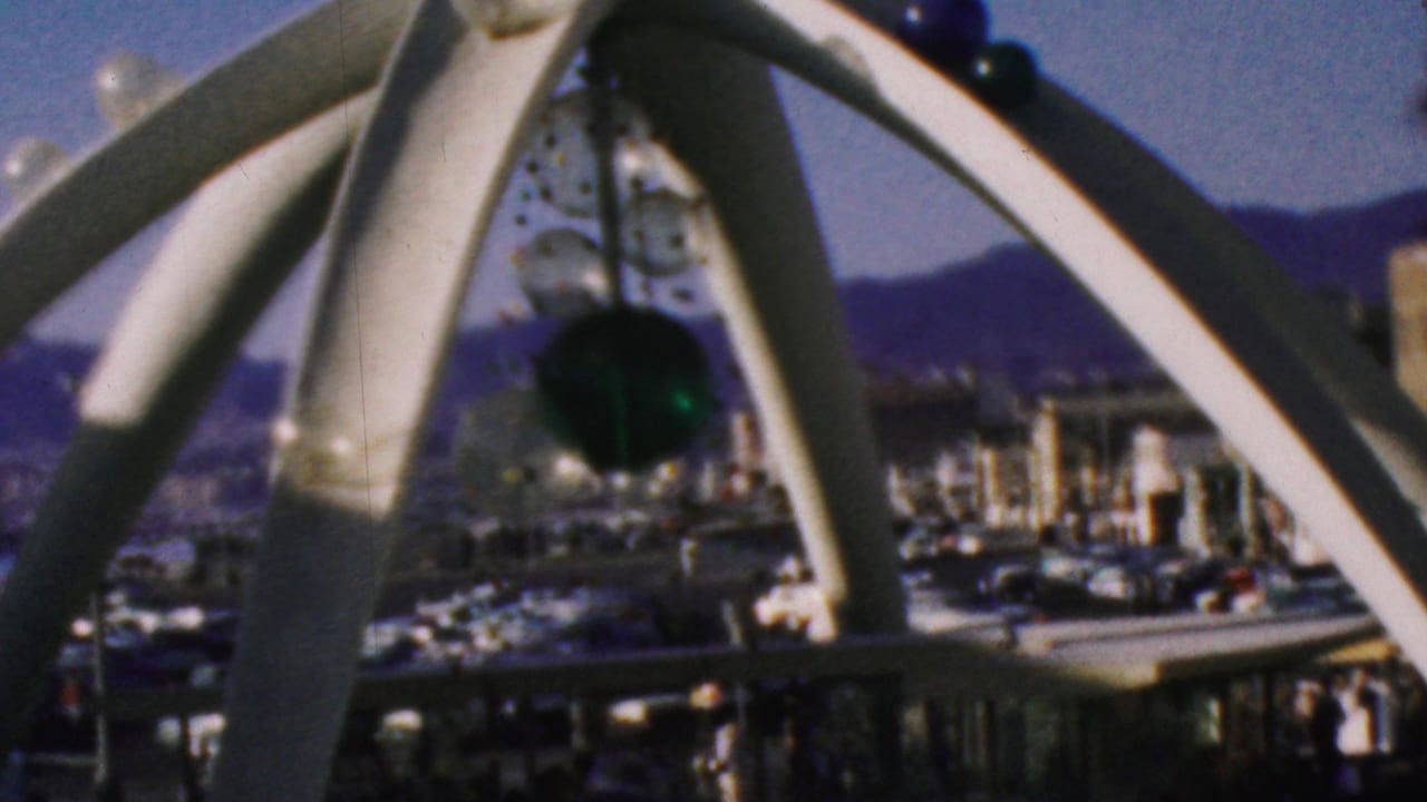 Visitor-shot 1964 Anaheim Disneyland footage showing a white arched outdoor sculpture with suspended green and clear glass elements against blue sky and distant park structures.