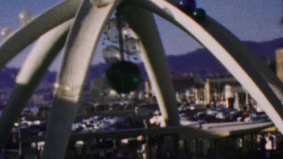 Visitor-shot 1964 Anaheim Disneyland footage showing a white arched outdoor sculpture with suspended green and clear glass elements against blue sky and distant park structures.