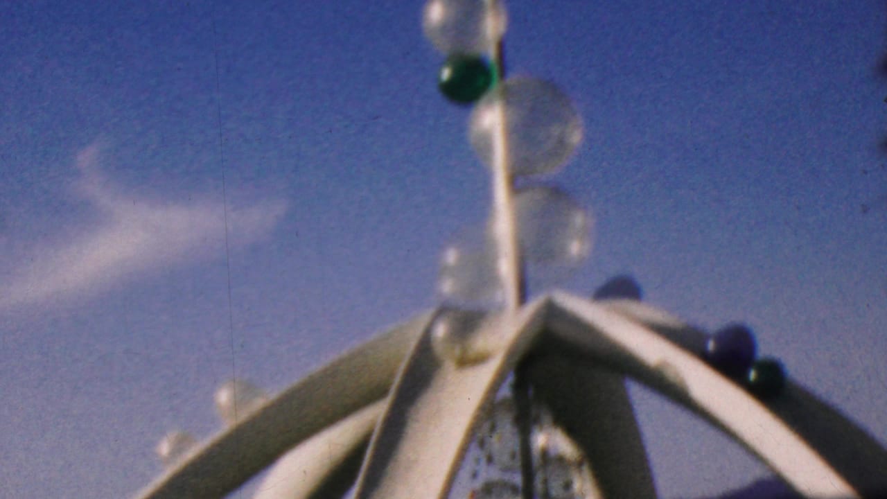 1964 Disneyland home-movie frame showing a decorative pole with green and clear glass orbs rising above white arched attraction structures under a blue sky.