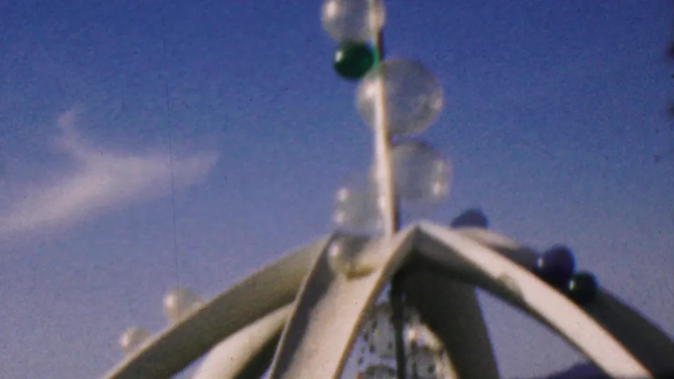 1964 Disneyland home-movie frame showing a decorative pole with green and clear glass orbs rising above white arched attraction structures under a blue sky.