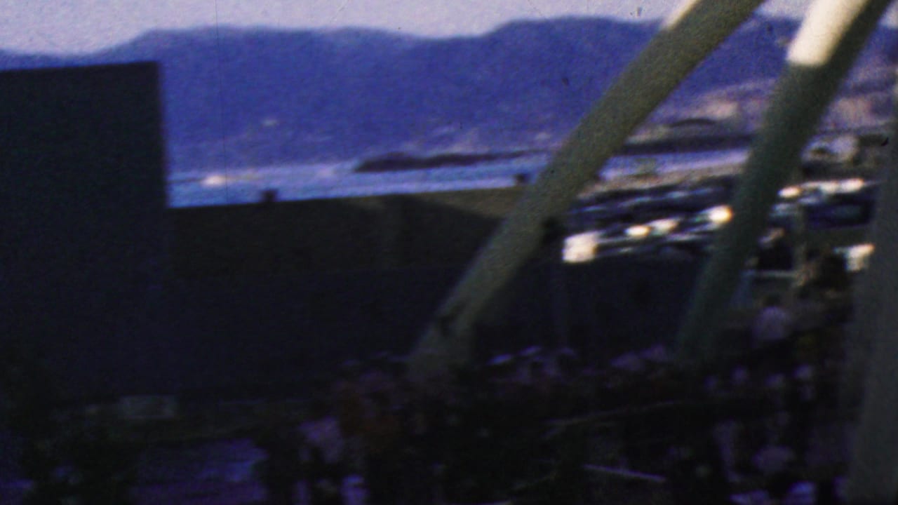 Crowded waterfront area seen through a white arched structure with mountains behind.