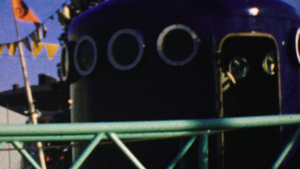 1964 Disneyland frame showing a dark attraction vehicle or structure with circular portholes, pennant flags above, and a green railing across the foreground.