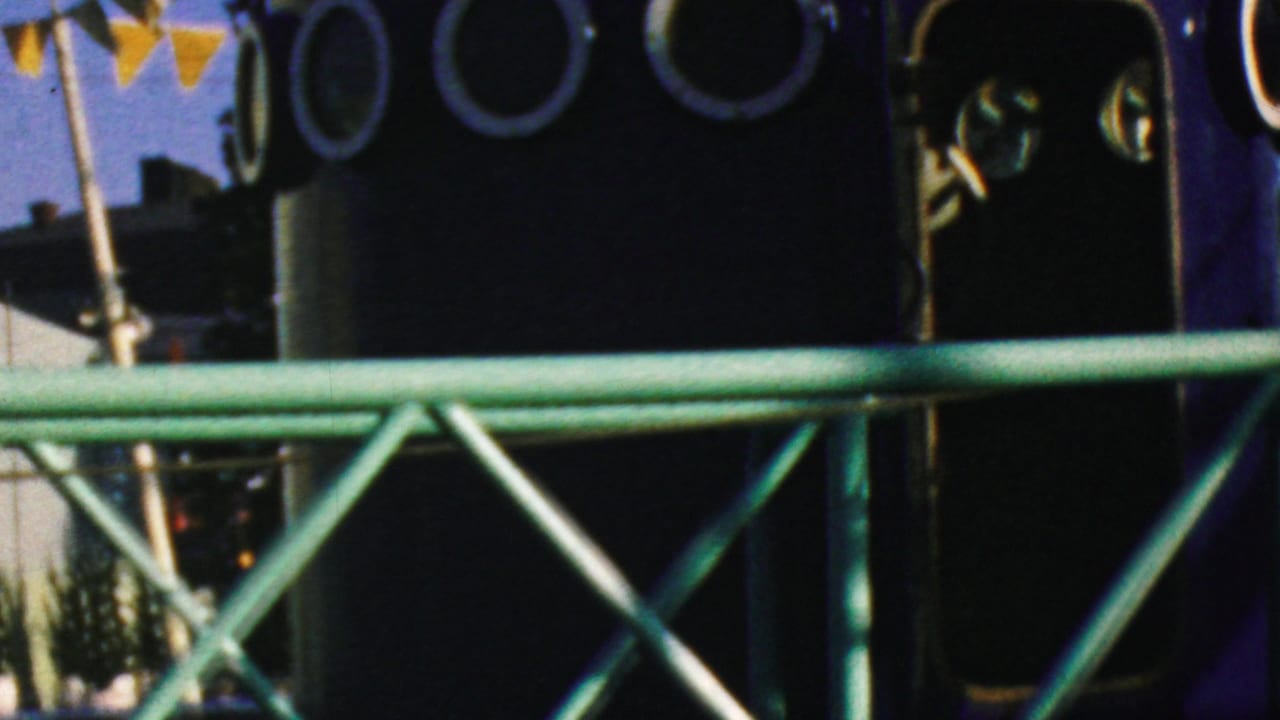 1964 Disneyland home-movie frame showing green metal railings crossing a dark blue rounded ride structure with circular porthole windows and triangular bunting overhead.