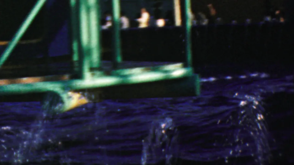 Visitor-shot 8mm frame from Disneyland in 1964 showing green railings, a dark ride structure, rippling water, and distant park visitors beyond the attraction area.