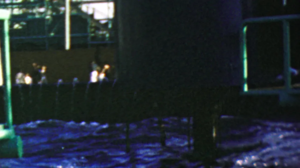 1964 Disneyland home-movie frame with a green vertical support, dark ride wall, blue churning water, background railings, and small indistinct visitors in the distance.