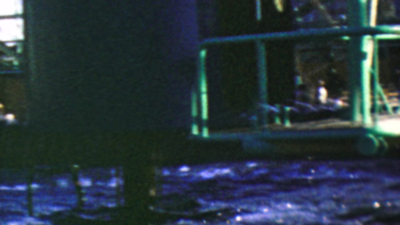 1964 Anaheim Disneyland home-movie frame showing a green metal water-ride structure over dark blue rippling water, with distant guests partly visible in the background.