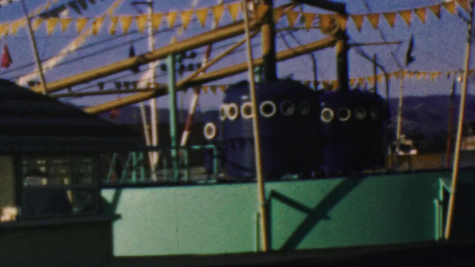 1964 Disneyland home-movie frame showing yellow pennant banners above a green water-ride structure, with posts, railings, and distant park elements visible.