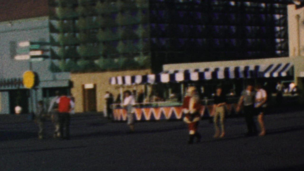 1964 Disneyland visitor view showing a plaza with striped awnings, guests walking and standing, and a Santa-costumed figure in front of a modern attraction facade.