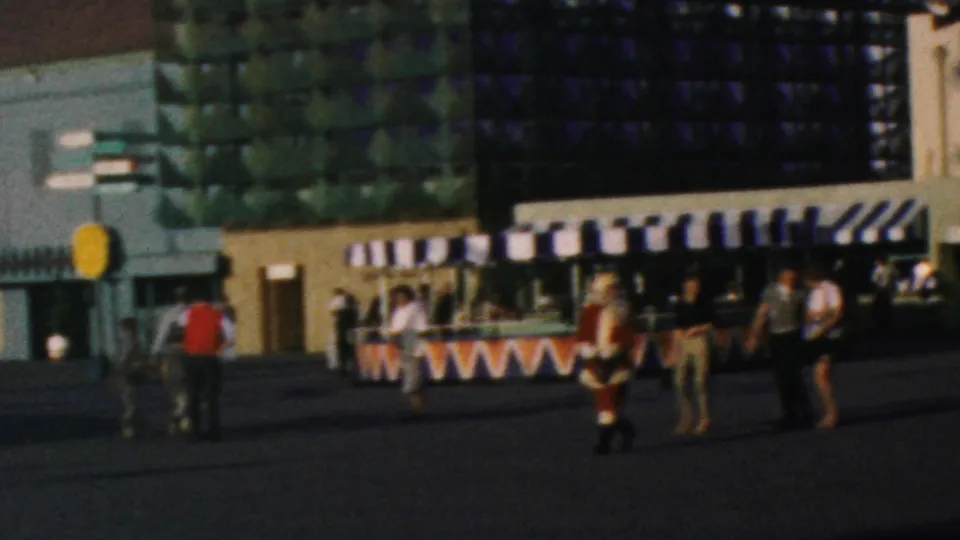 1964 Disneyland visitor view showing a plaza with striped awnings, guests walking and standing, and a Santa-costumed figure in front of a modern attraction facade.