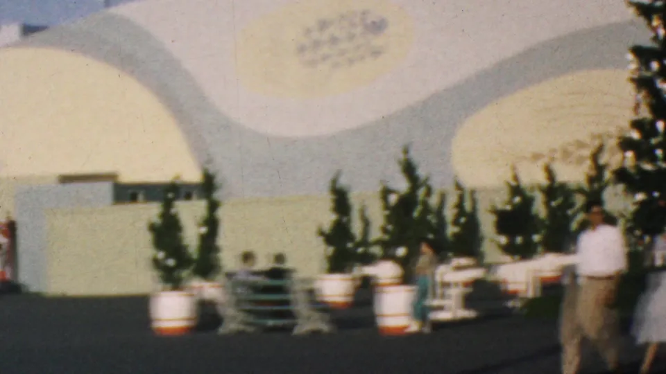 1964 Disneyland plaza view showing a curved white building wall, potted evergreens, benches, barrel planters, and visitors standing or walking nearby.