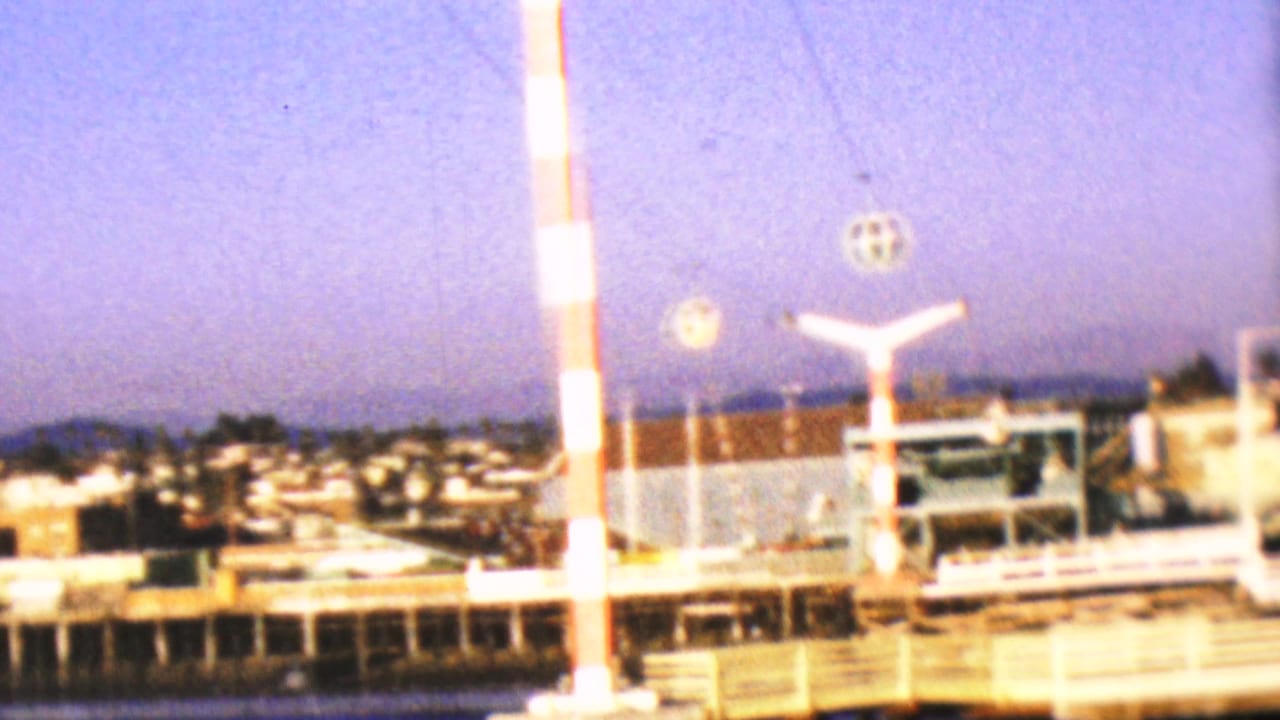1964 8mm home-movie frame from the Anaheim, California clip showing a red-and-white striped pole, bright waterfront structures, and distant low-rise buildings across the water.