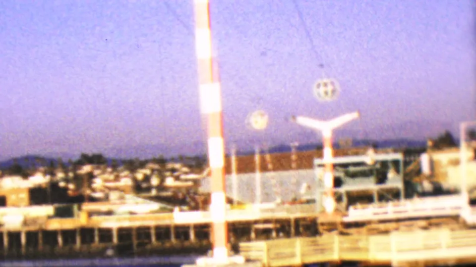 1964 8mm home-movie frame from the Anaheim, California clip showing a red-and-white striped pole, bright waterfront structures, and distant low-rise buildings across the water.