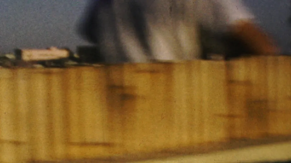 Soft low-light 8mm frame from the Anaheim Disneyland clip showing a tan foreground barrier or boat edge, blurred background structures, and warm night lighting.