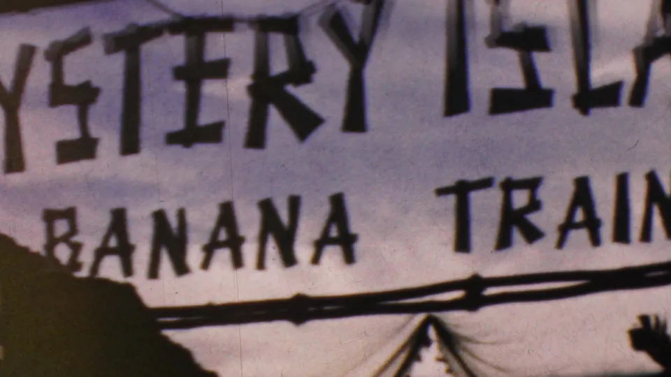 Visitor-shot 1964 Anaheim frame showing a cropped Mystery Island sign with Banana Train text, silhouetted foreground edges, and visible 8mm softness.