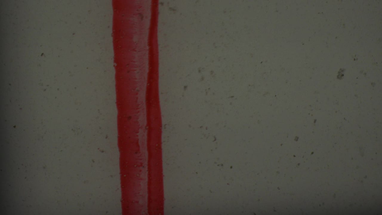 Close-up of a red stamped vertical mark with a rounded lower end on a pale textured surface, from supplied 1965 Los Angeles 8mm footage.