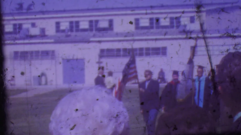 In Los Angeles 1965 home-movie footage, a small group in graduation-style clothing stands outdoors with American flags and a pale banner in front of a large school-like building.