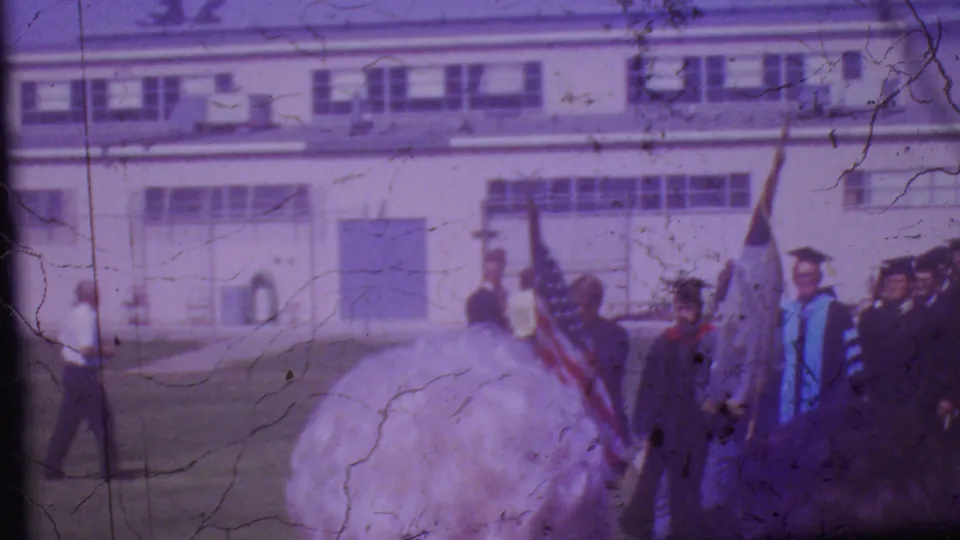The 1965 Los Angeles frame shows ceremony participants gathered outside a large building, with American flags, a pale banner, and several visible faces in soft, scratched 8mm color.
