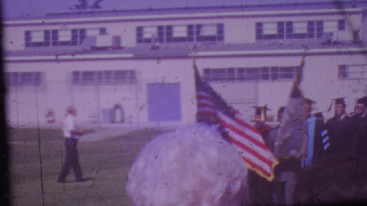 1965 Los Angeles home-movie frame showing graduates in caps and gowns carrying flags across a grassy field outside a large institutional building.