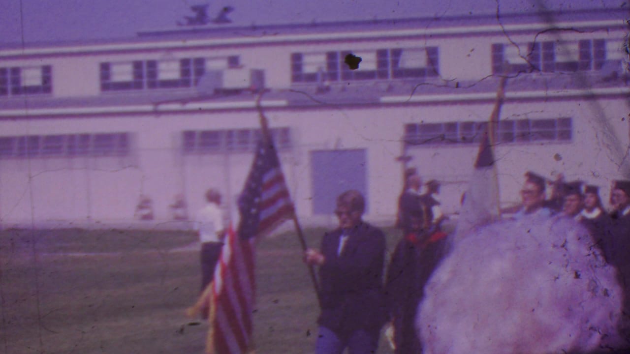 1965 Los Angeles graduation procession frame with an American flag, graduates in dark robes, and adult onlookers near a school-like building.