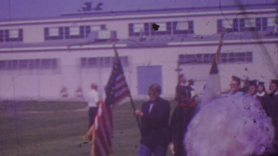 1965 Los Angeles graduation procession frame with an American flag, graduates in dark robes, and adult onlookers near a school-like building.
