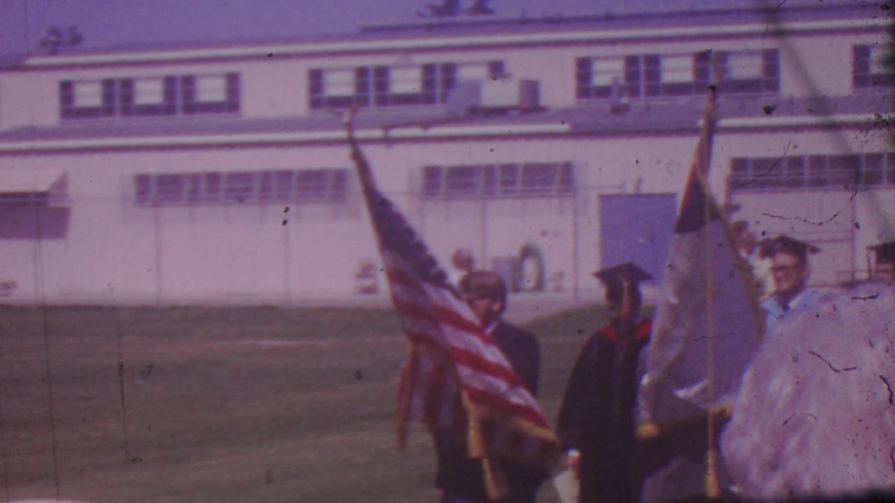 Two flag bearers and a graduate walk across the field in front of the building.