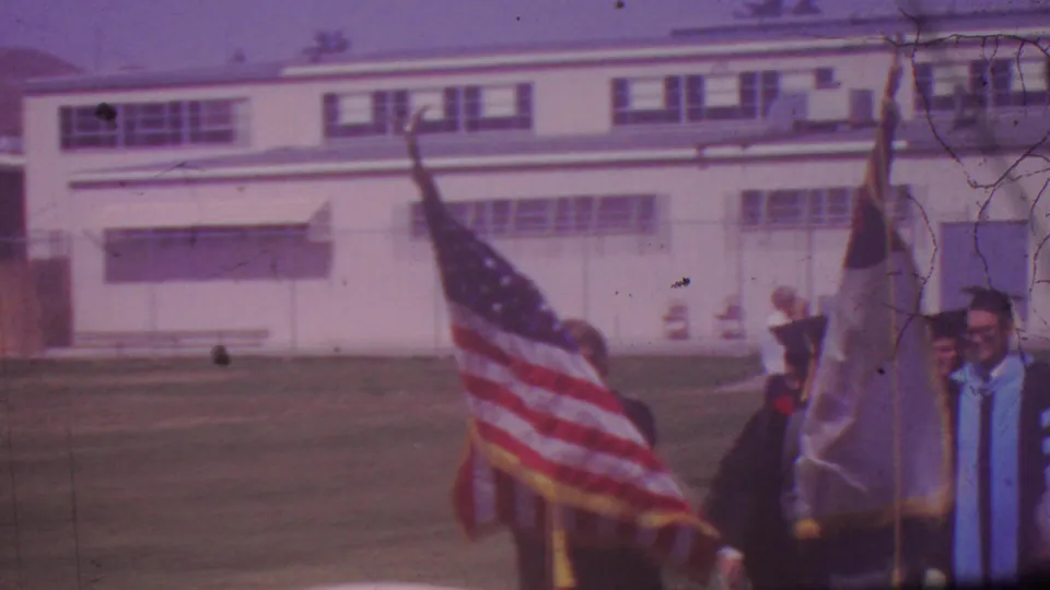 1965 Los Angeles graduation procession frame with flags filling the foreground and robed participants partially visible beside a large school-like building.