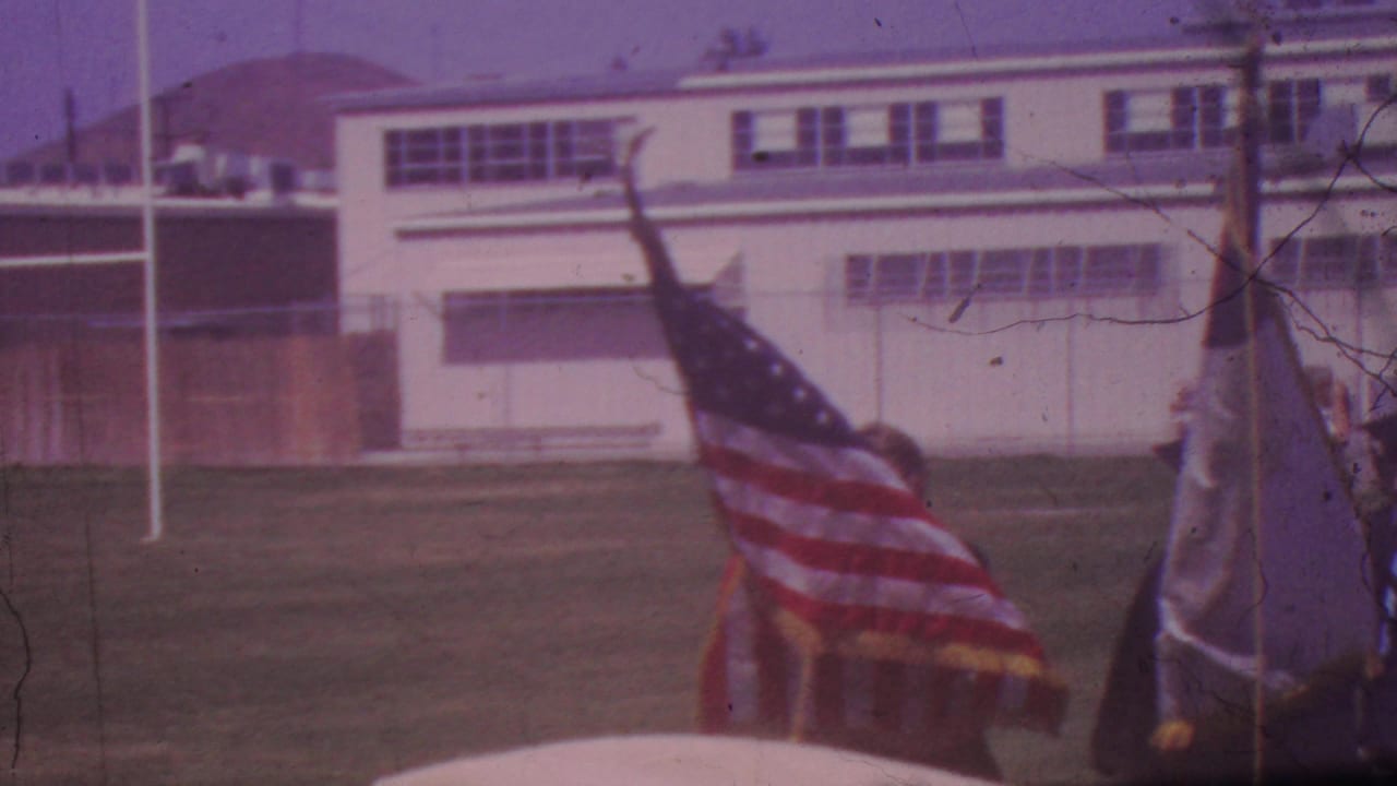 1965 Los Angeles high school graduation procession with an American flag, a white school building, and graduates crossing a grassy field.