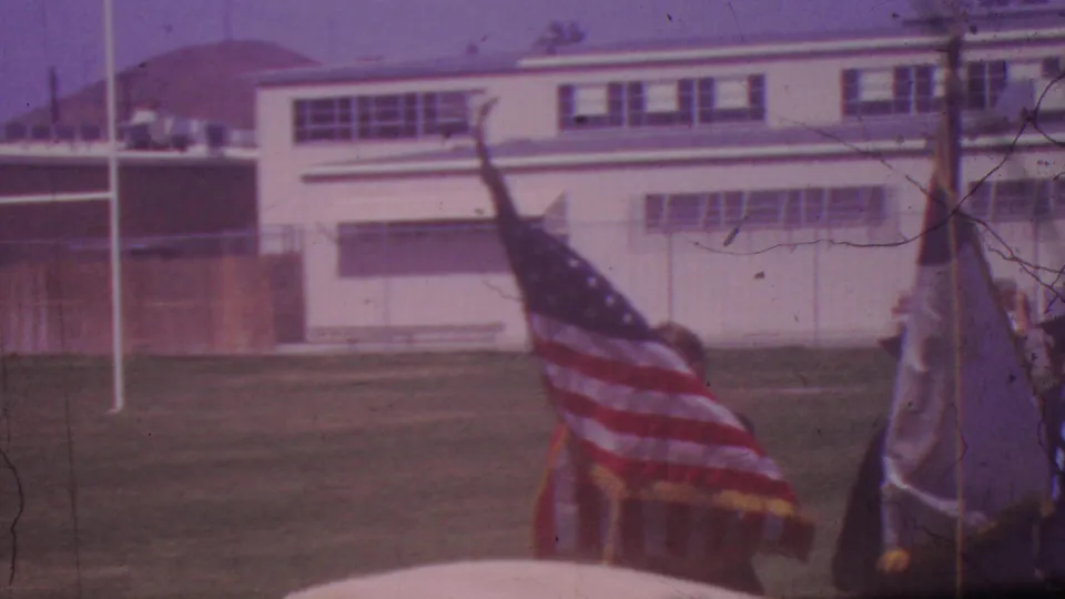 1965 Los Angeles high school graduation procession with an American flag, a white school building, and graduates crossing a grassy field.