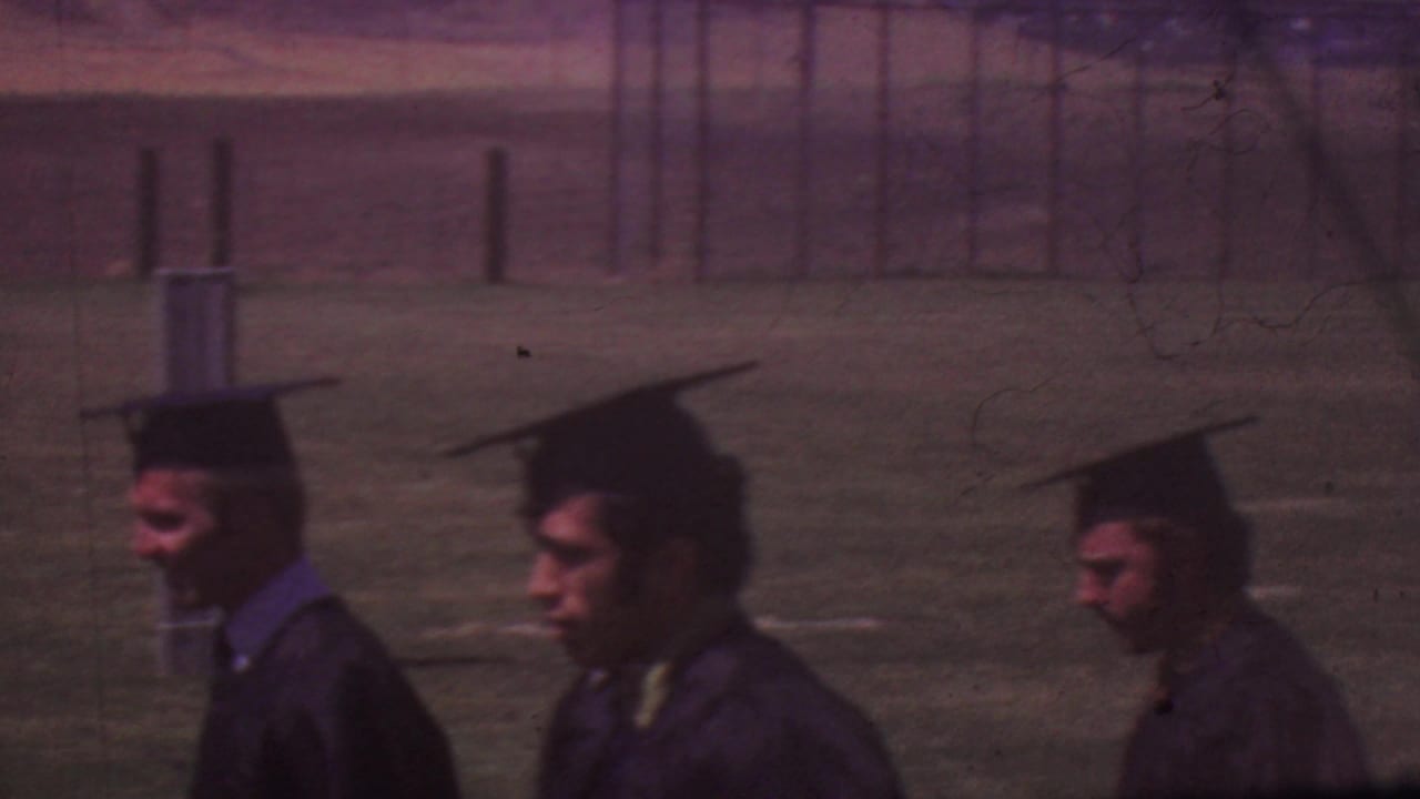 Graduates in black mortarboards and gowns process across an outdoor school field during the Los Angeles graduation ceremony.