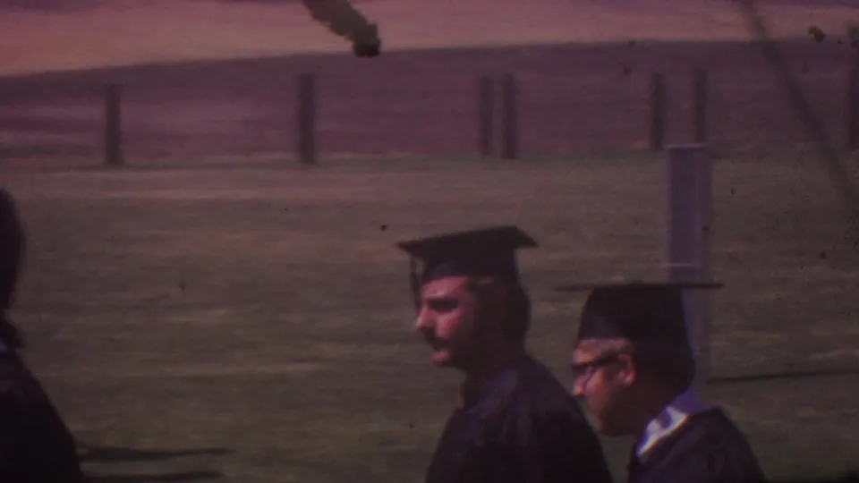 Two male-presenting graduates in black caps and gowns pass close to the camera during the outdoor 1965 graduation procession.