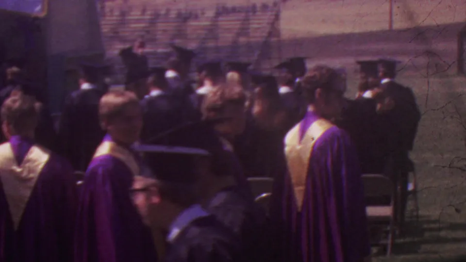 Faculty or ceremony officials in academic robes and graduates gather near folding chairs during the outdoor Los Angeles high school graduation.