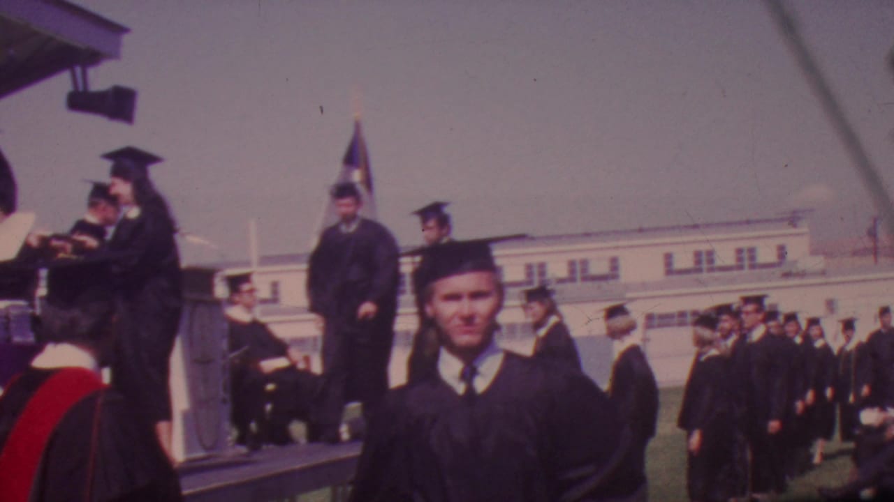 1965 Los Angeles graduation footage showing black-gowned students near a small outdoor stage with a white institutional building in the background.