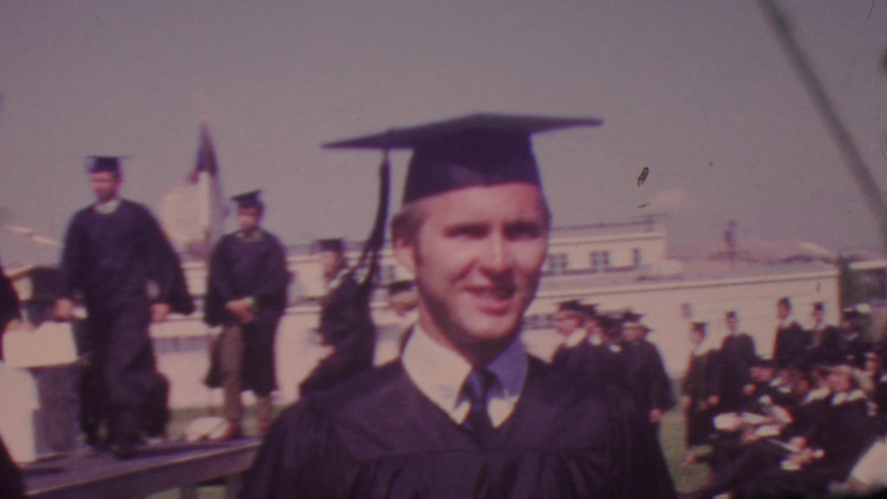 A young male graduate in black cap and gown smiles toward the handheld camera during the Los Angeles graduation ceremony, with other graduates and spectators behind him.